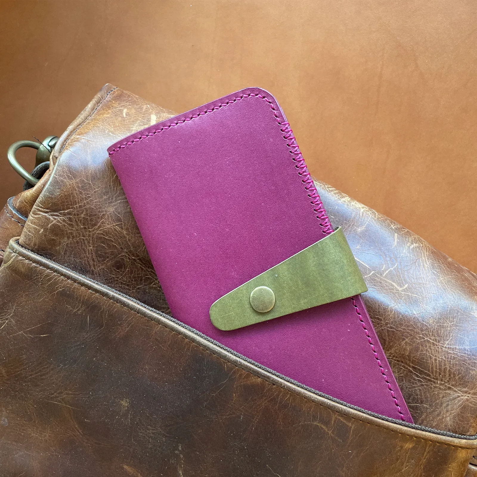 A purple long wallet with an olive snap strap tucked into a worn leather bag
