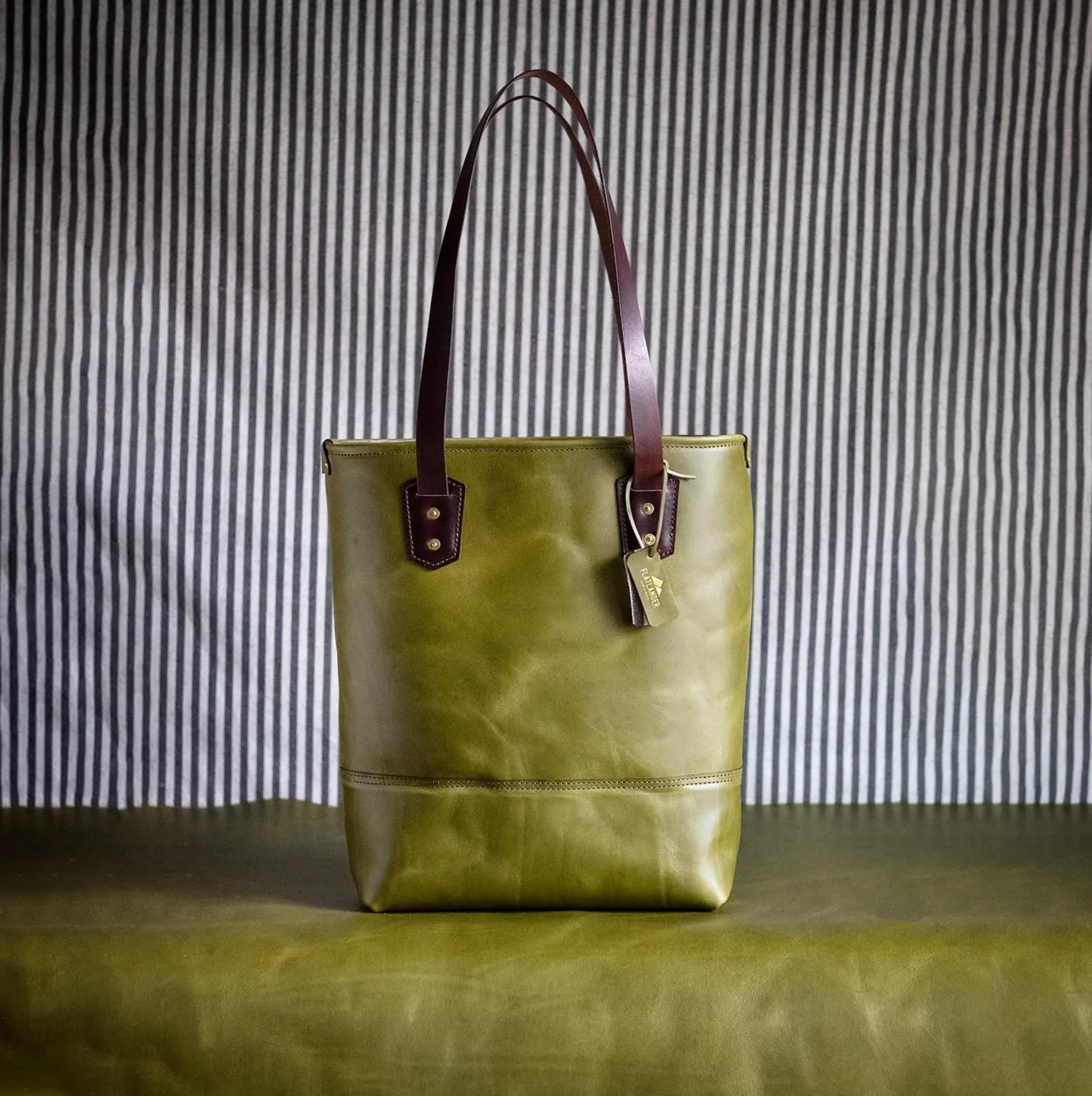 An olive leather tote with dark leather handles, photographed against ticking-stripe fabric