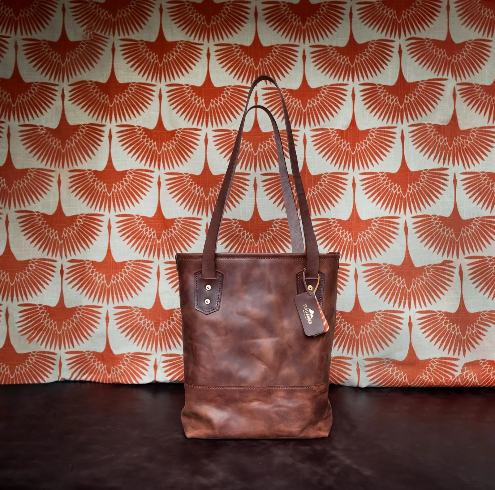 A brown leather tote against a wallpaper of printed orange wings