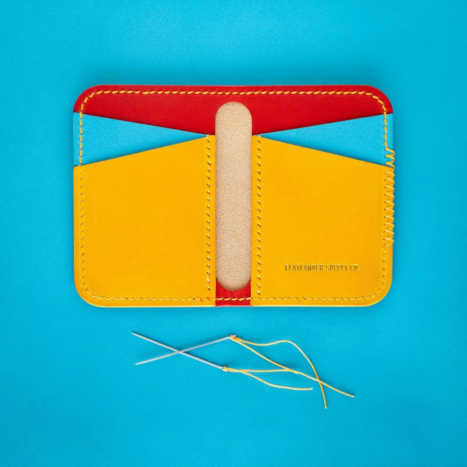 A card wallet open flat on a cyan leather hide in yellow, red and blue panels, with loose waxed thread beside it