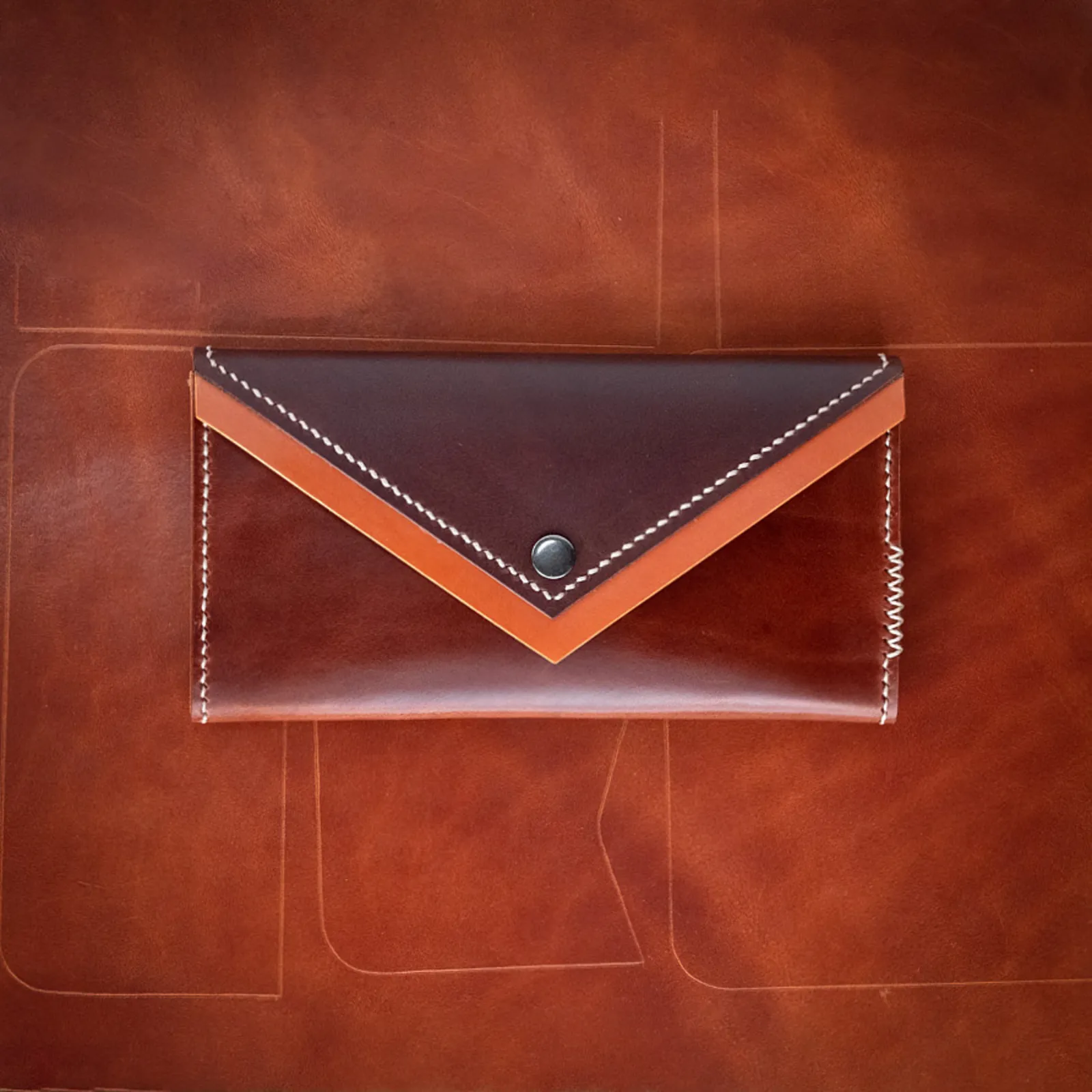 An oxblood and tan envelope clutch photographed on a sheet of the same leather, showing the cutting lines it came from