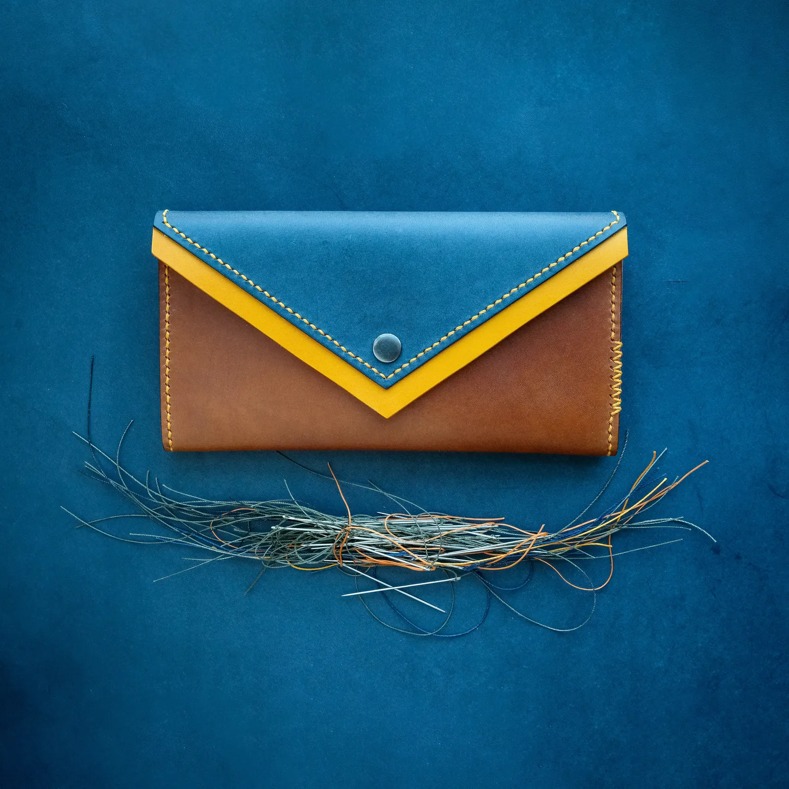 An envelope clutch in tan and blue leather with a V-shaped flap and a brass snap, on a blue backdrop
