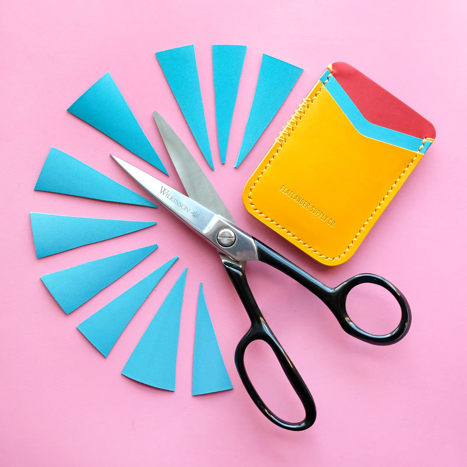 Cyan leather offcuts fanned into a starburst around a pair of black shears, with a yellow card sleeve, on a pink leather hide