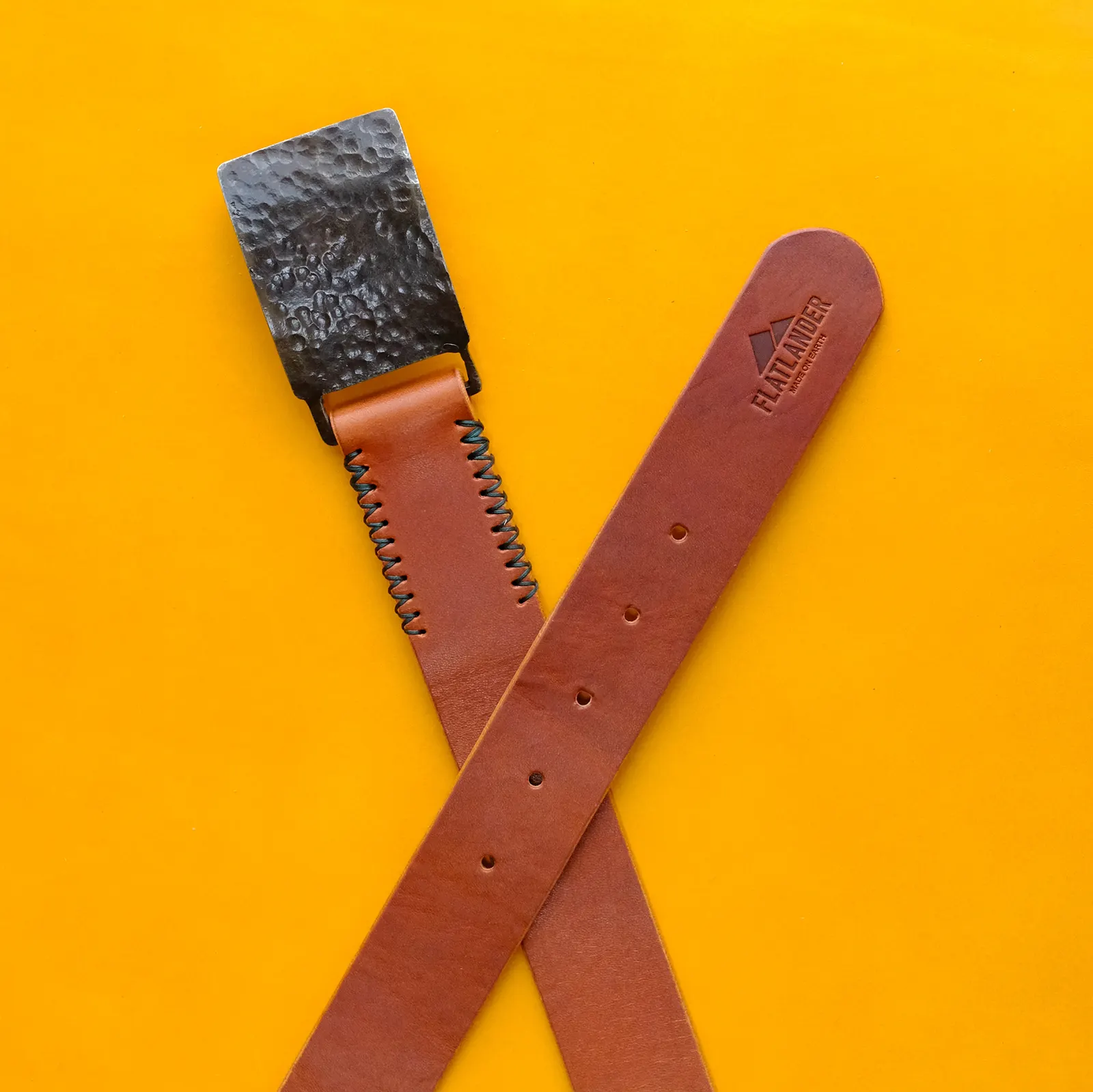Two hand-stitched leather belts crossed on a yellow leather hide beside a hammered buckle