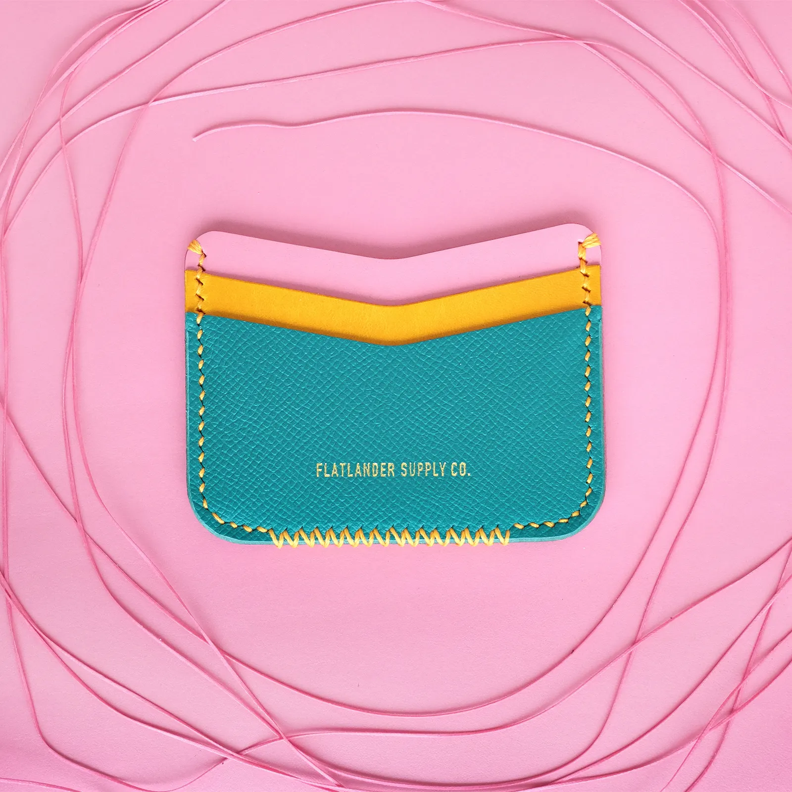 A teal card sleeve with yellow contrast stitching, centered in a nest of coiled pink cord on a pink leather hide